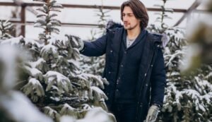 "Man wearing a denim jacket layered over winter attire, standing outdoors in a snowy park."