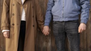 "Man wearing a denim jacket layered under a long overcoat, standing outdoors."