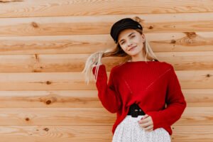 Blonde woman in a red sweater dress posing outdoors near a wooden house, styled for casual street fashion