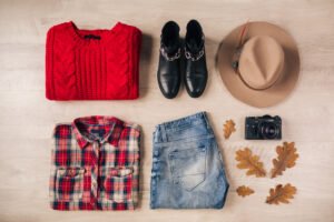 Flat lay of a red knitted sweater with boots, denim jeans, checkered shirt, hat, and vintage camera for fall fashion